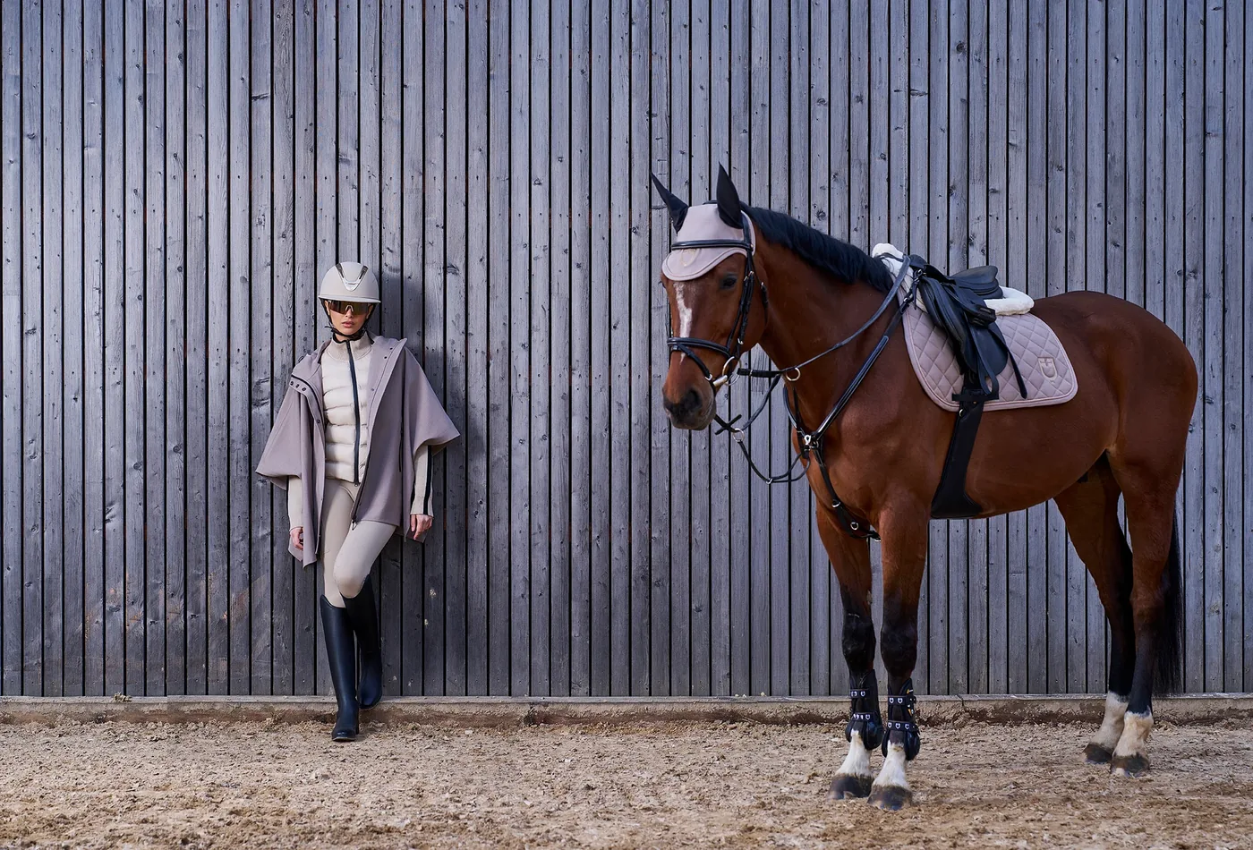 Braunes Pferd mit heller Schabracke, Fliegenhaube und schwarzen Gamaschen neben einer Reiterin im Poncho vor einer Holzwand
