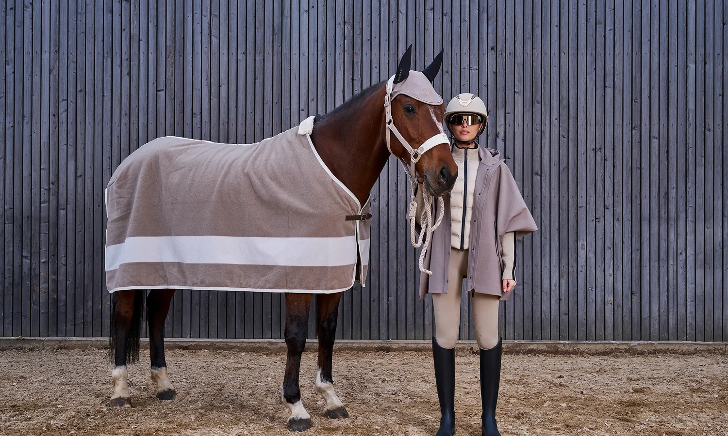 Reiterin im hellen Equestro-Outfit mit Reithelm neben einem braunen Pferd mit abgestimmter Abschwitzdecke und Fliegenhaube