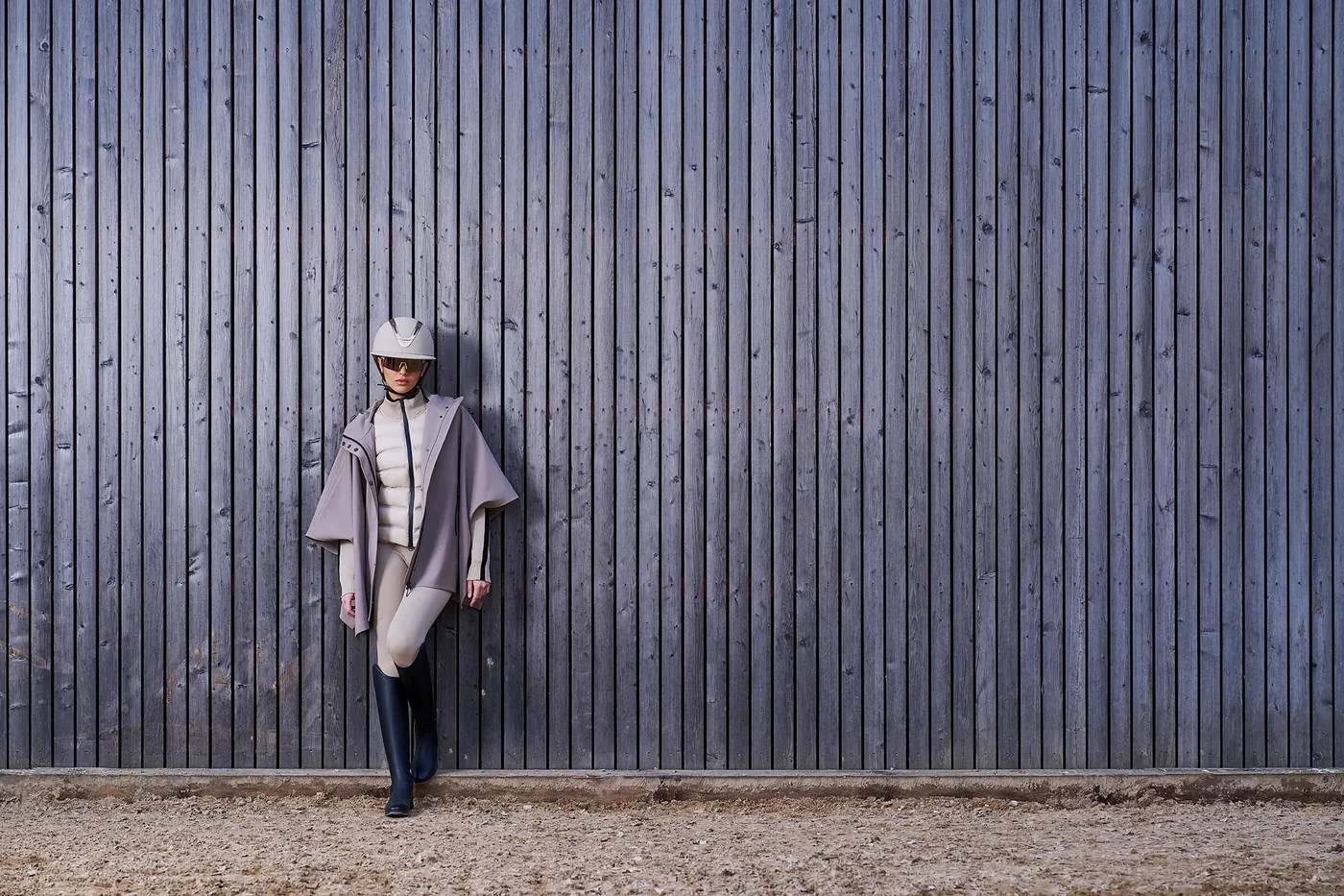 Reiterin mit Helm, Poncho, heller Reithose und schwarzen Reitstiefeln lehnt an einer Holzwand