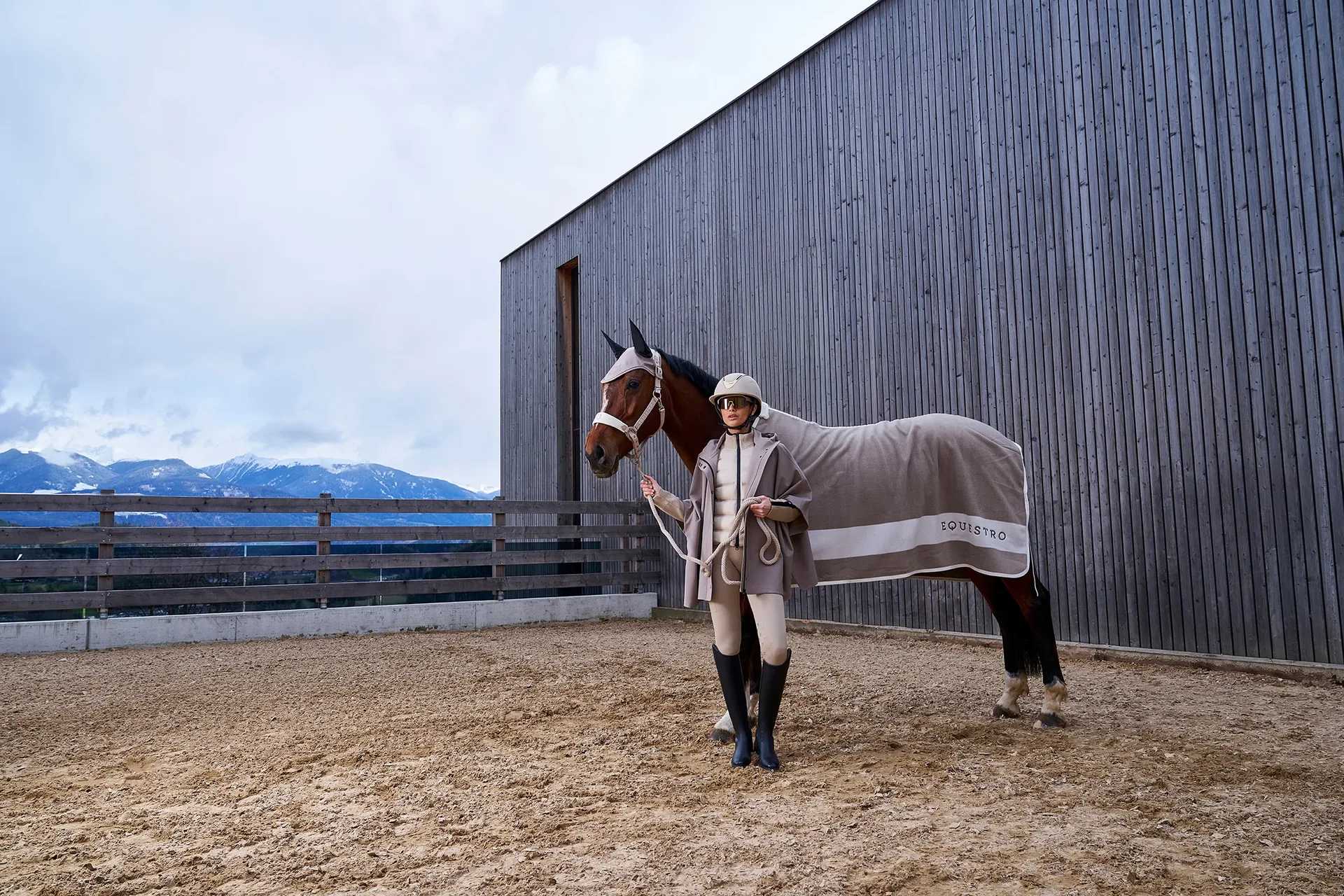 Ruiter met helm en poncho leidt haar paard in een grijze Equestro zweetdeken over de rijbaan voor een houten hal